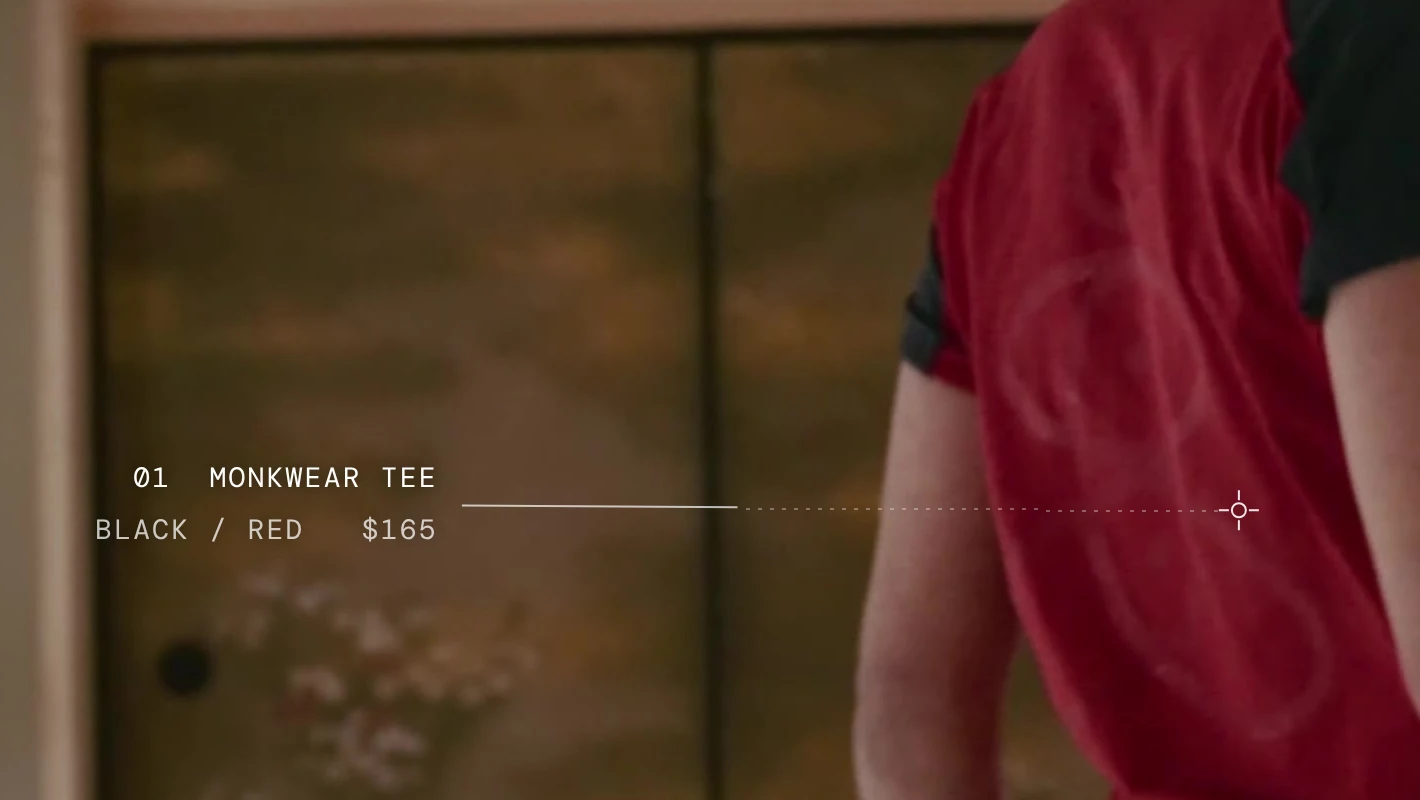 The tee’s credit up close, from the home page: on the left, 01 Monkwear Tee and Black / Red $165 in white mono type before the gold screens; a leader runs right, solid in the open and dotted where it crosses his arm, to a small ring with four ticks on the red back of his tee